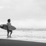 Surfer standing on a beach with a surfboard looking at the waves on an overcast day