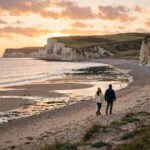 A couple walking hand in hand along a quiet beach at sunset on a romantic UK getaway.
